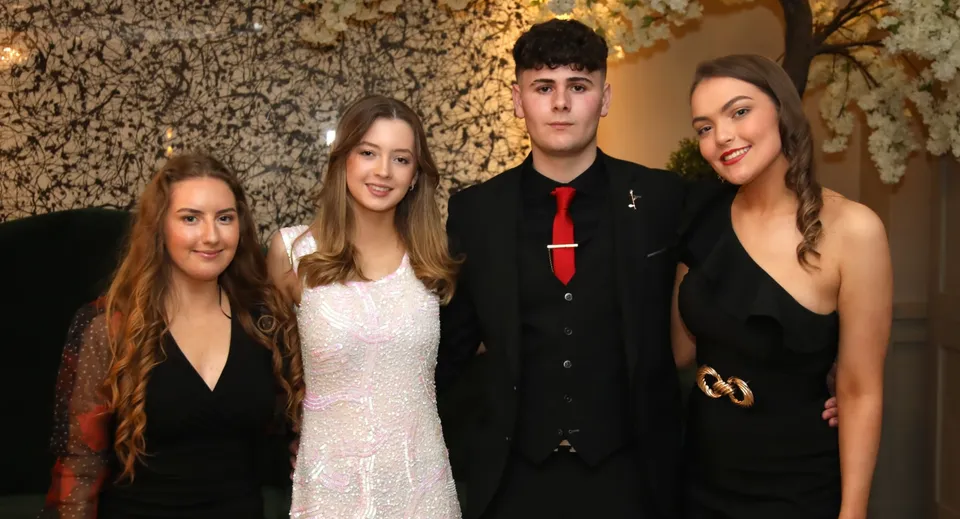 Quattro studenti della Royal School posano in abiti formali davanti a una parete in pietra strutturata.