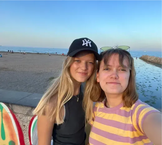 Zwei Mädchen lächeln für ein Selfie am Strand in Spanien, im Hintergrund das Meer