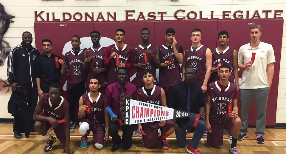 La squadra di basket del Kildonan East Collegiate posa insieme a un trofeo davanti a un cartello della scuola.