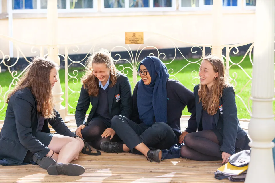 Quattro studentesse della Torquay Girls Grammar School sedute insieme su una panchina davanti a un cancello decorativo in metallo.