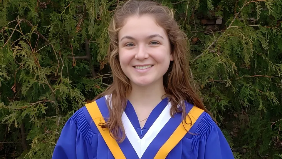 Una giovane donna in toga da diploma blu e gialla si trova davanti a uno sfondo verde nel campus del Collège Sacré-Coeur.