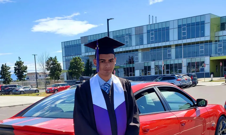 Uno studente della Royal Bay Secondary School con la toga di diploma davanti a un'auto rossa di fronte a un edificio moderno.