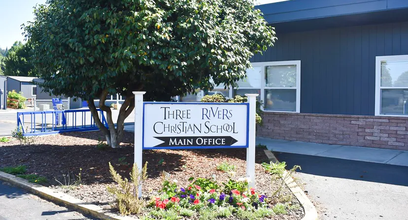 L'insegna della Three Rivers Christian School si trova in un'aiuola ben curata con un albero davanti a un edificio blu.