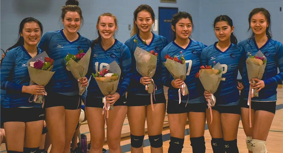 Un gruppo di giovani giocatrici di pallavolo in maglia blu tiene mazzi di fiori nella palestra della Port Moody Secondary School.