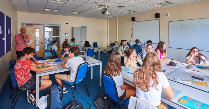 Aula del college con estudiantes en las mesas y el profesor de pie junto a la puerta