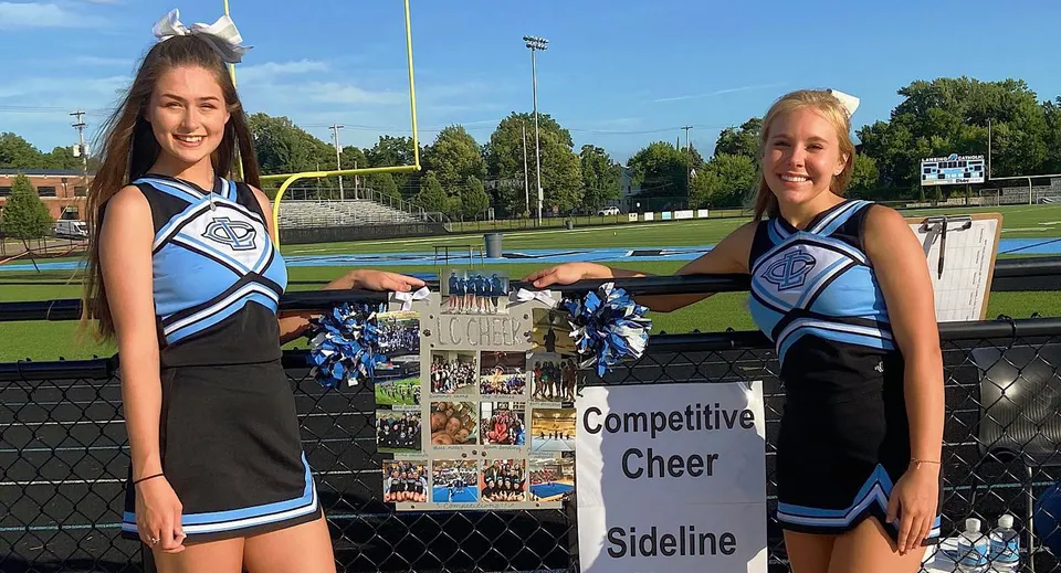 Due cheerleader in divisa blu e bianca si trovano a bordo campo della Lansing Catholic High School davanti a un cartello sportivo.
