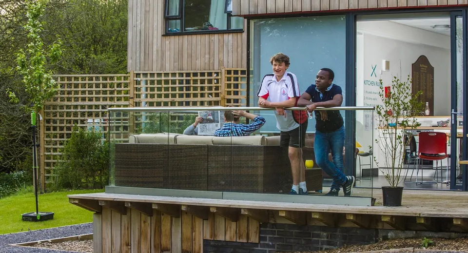 Due persone si trovano sulla terrazza in legno della Kingswood School davanti a una recinzione in legno e piante verdi.