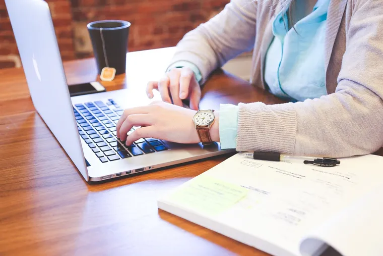 Person tippt auf einem Laptop neben Notizblock und Teetasse am Holztisch