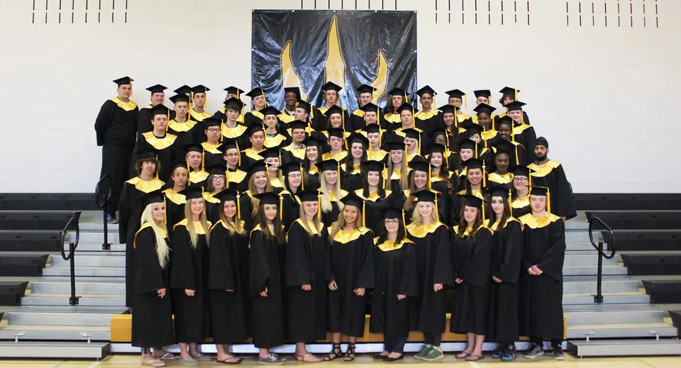 Un grande gruppo di studenti e studentesse della Drumheller Valley School indossa toghe nere da diploma davanti a striscioni giallo-blu.