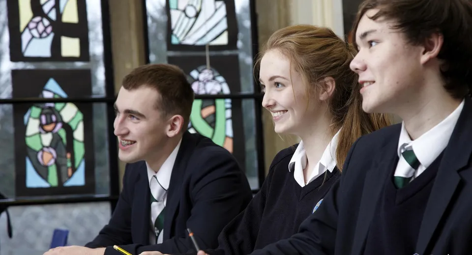 Tre studenti della Durham School sorridono davanti a una vetrata colorata.