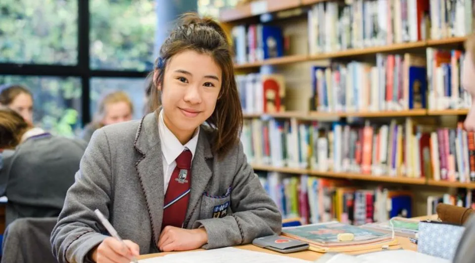 Una studentessa del Victoria College Belfast è seduta a un tavolo in biblioteca tra gli scaffali dei libri.