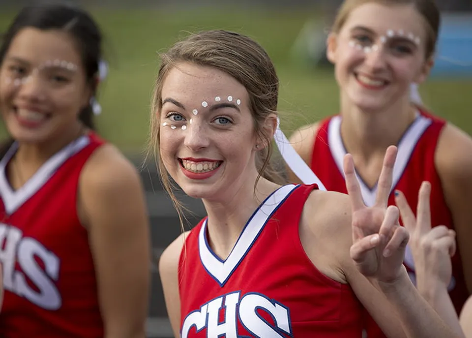 Tre studentesse in uniformi da cheerleading rosse fanno gesti entusiasti sul campo della Red Creek High School.
