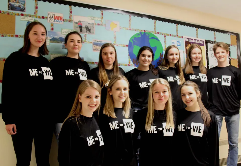 Un gruppo di studentesse con t-shirt nere ME + WE si trova davanti a una parete coperta di materiali didattici nella Corner Brook Regional High School.