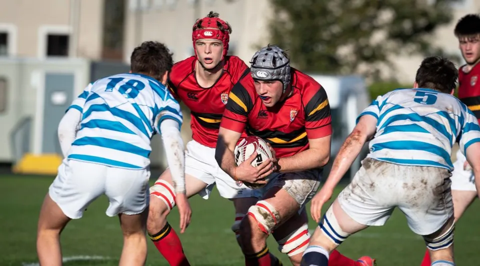 Studenti del Stewart’s Melville College giocano un'intensa partita di rugby su un campo erboso con edifici sullo sfondo.