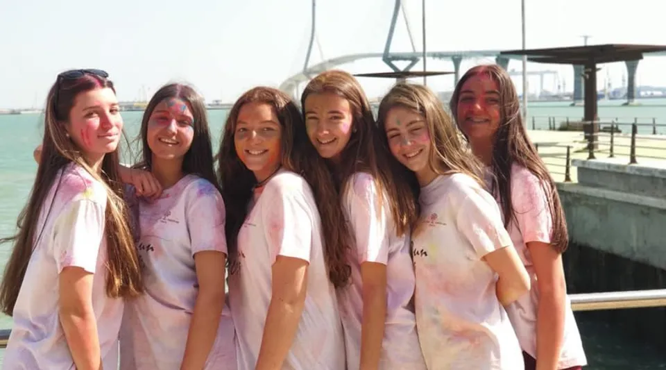 Un gruppo di studenti della San Vicente de Paul School indossa magliette uniformi rosa e bianche davanti a uno specchio d'acqua con un ponte visibile.