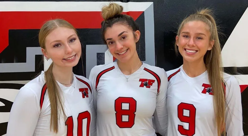 Tre studentesse della Terry Fox Secondary School indossano maglie sportive bianche con numeri rossi davanti a uno sfondo rosso e nero.
