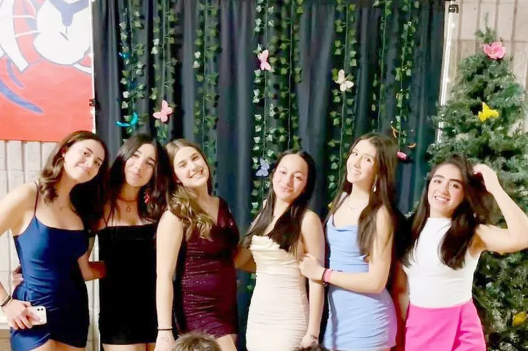 Seis jóvenes sonrientes posan juntas vestidas de fiesta delante de un fondo oscuro decorado