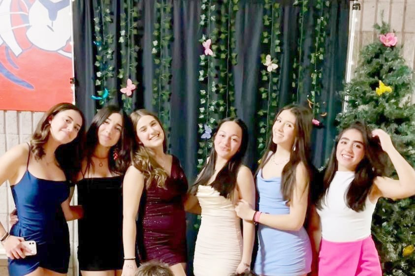 Seis jóvenes sonrientes posan juntas vestidas de fiesta delante de un fondo oscuro decorado
