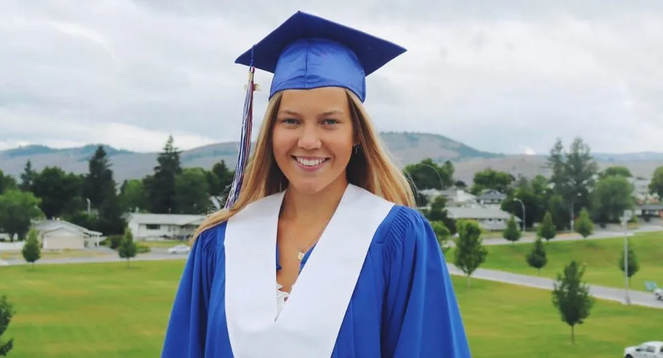 Una giovane donna con la toga blu da diploma si trova davanti alle montagne nel campus della Vernon Secondary School.