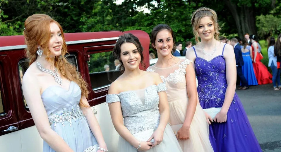 Quattro studentesse della Sacred Heart Secondary School Clonakilty in abito da sera stanno davanti a un'auto d'epoca rossa sotto gli alberi.