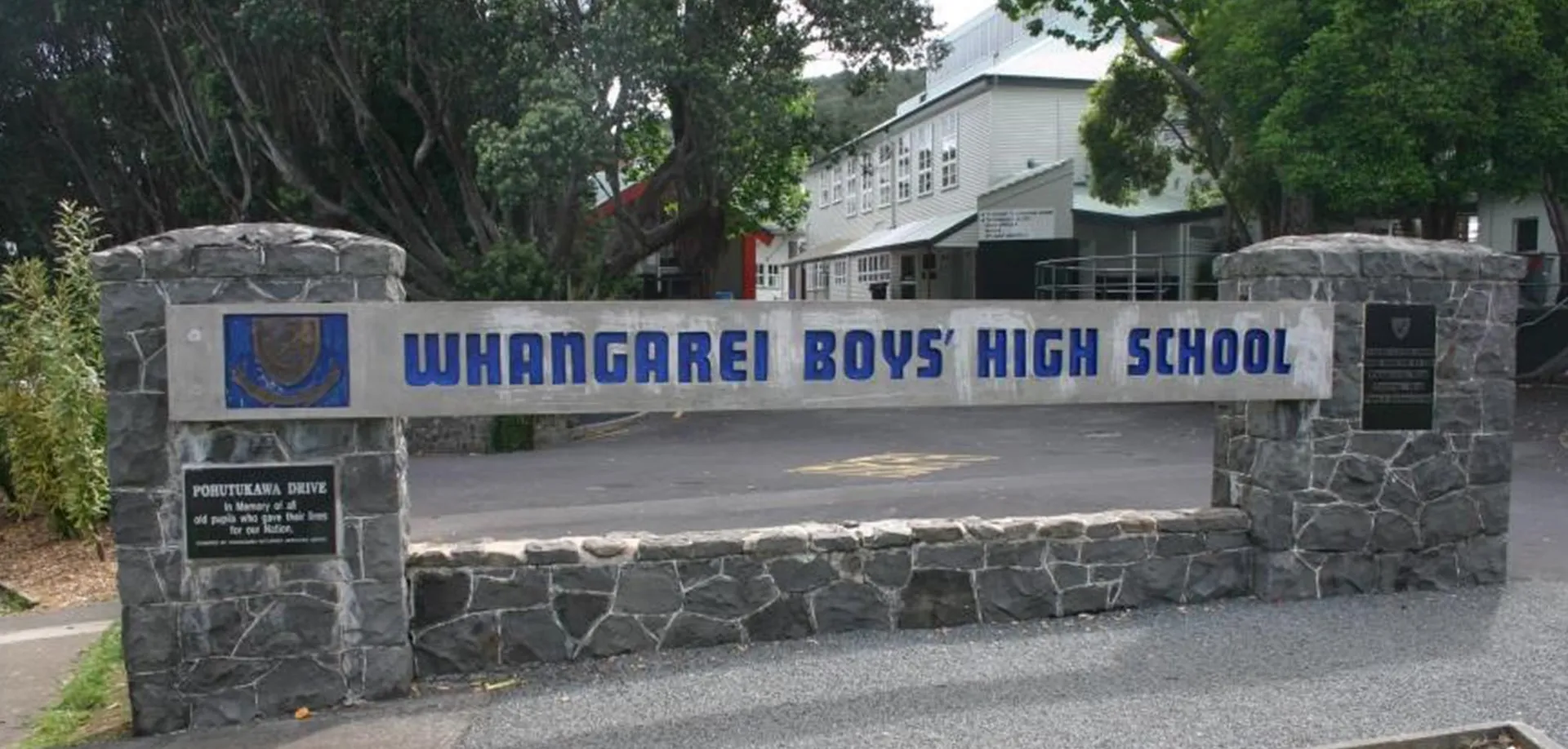 L'ingresso della Whangarei Boys’ High School con muro di pietra, insegna e un edificio sullo sfondo circondato da aree verdi.