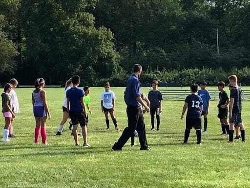 Estudiantes de la Saddle River Day School se reúnen con un entrenador en un campo de césped.