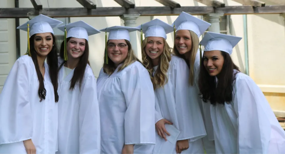 Un gruppo di giovani donne in abito da diploma bianco si trova nell'edificio scolastico della Academy of Notre Dame.
