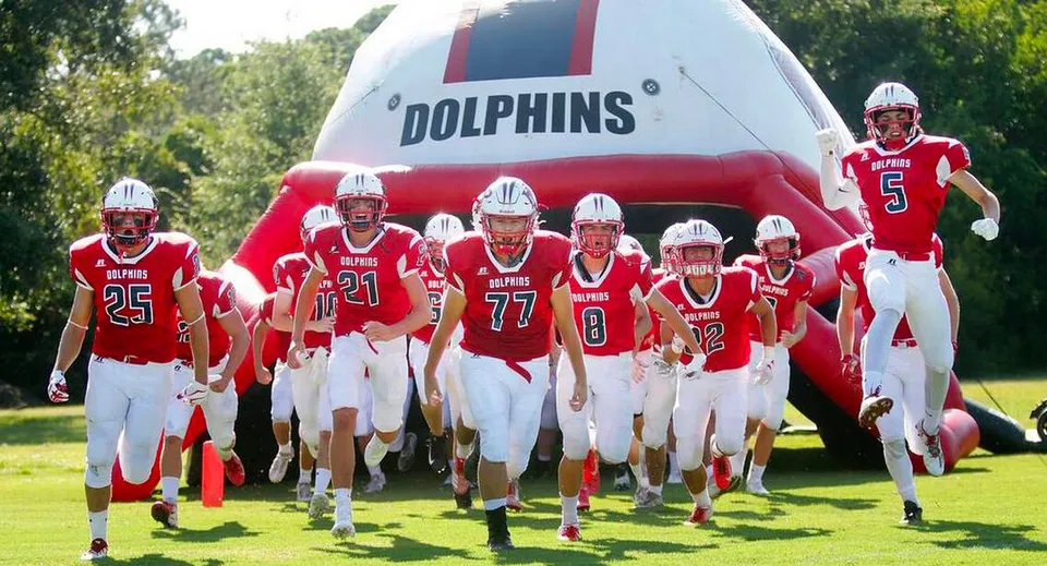 Giovani giocatori di football della Hilton Head Preparatory School in maglia rossa davanti a un grande logo gonfiabile dei Dolphins sul campo sportivo.