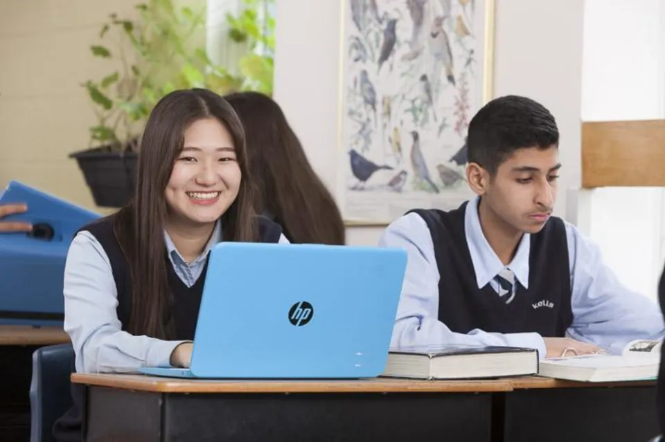 Due studenti della Kells Academy in divisa scolastica siedono in classe, mentre una studentessa lavora al computer portatile blu.