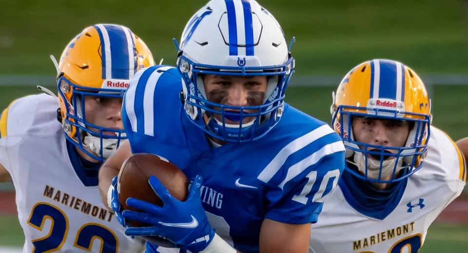 Tre giocatori di football si trovano sul campo da gioco della Bishop Mahoney High School con maglie blu e gialle.