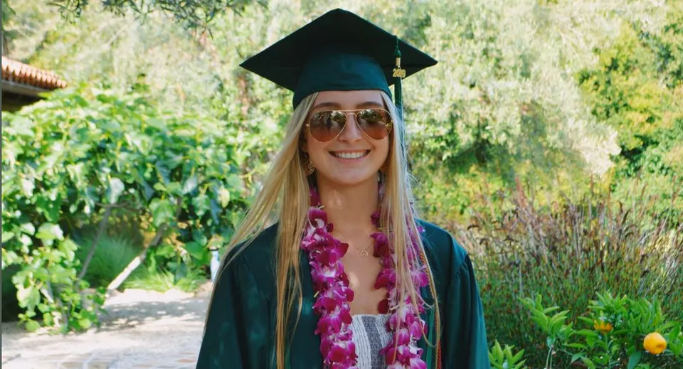 Una giovane donna in abito da diploma è in piedi nel verde del campus del Santa Barbara Unified School District.