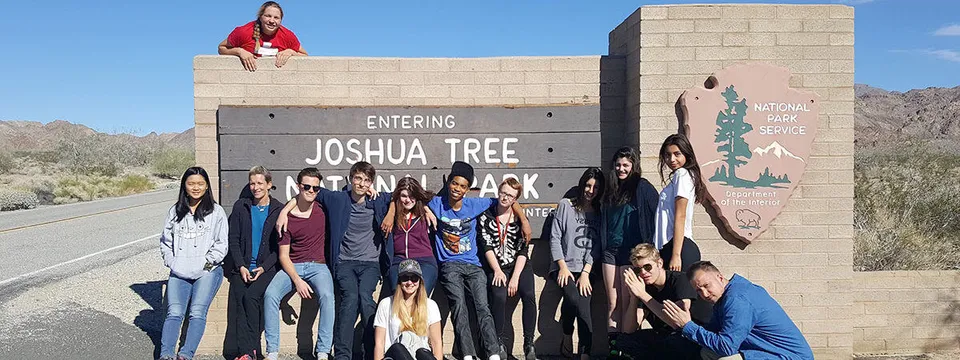 Studenti e studentesse della The Orme School si trovano davanti al cartello d'ingresso del Joshua Tree National Park in un paesaggio desertico.