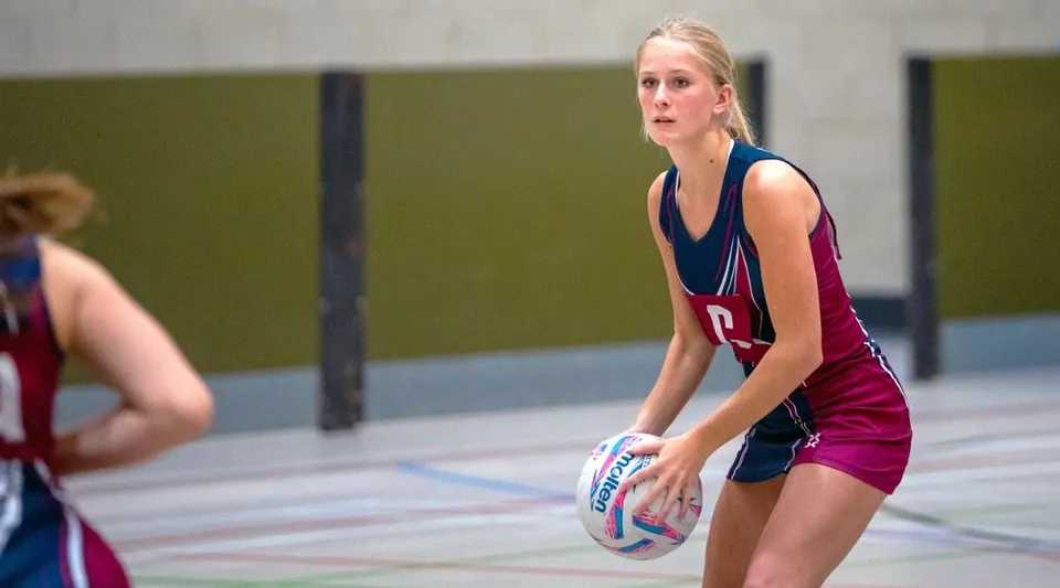 Una studentessa della Norwich School tiene un pallone da pallavolo sul campo sportivo con una parete verde sullo sfondo.