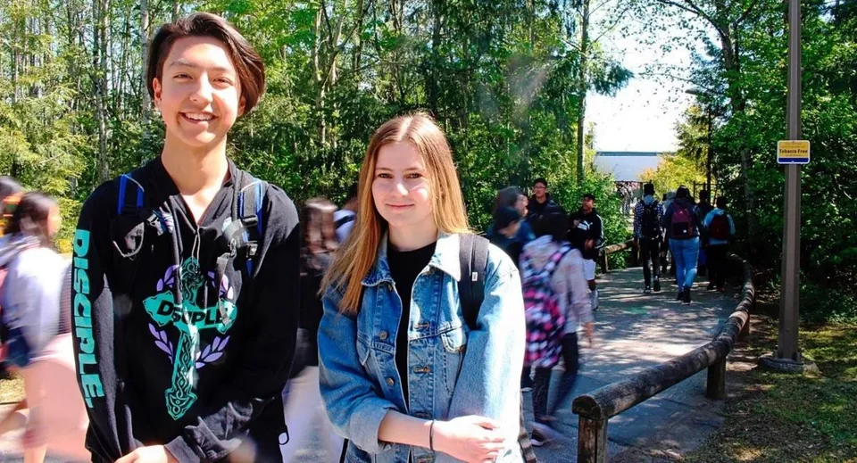 Due studentesse sono all'esterno nel campus della Fraser Heights Secondary School davanti ad alberi e a una folla di persone.