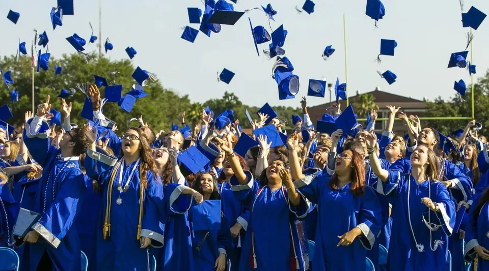 Un grande gruppo di diplomati delle Marion County Public Schools lancia in aria i tocchi blu davanti a uno sfondo di alberi.