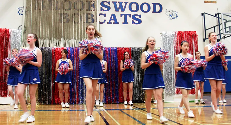 Studenti e studentesse della Brookswood Secondary School si esibiscono nel cheerleading su un campo da basket con l'insegna Swood Cats sullo sfondo.