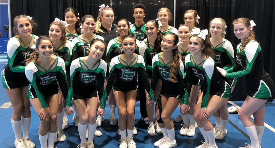 Un gruppo di giovani ginnaste della Lord Tweedsmuir Secondary School posa insieme in divisa verde e nera su un palco.