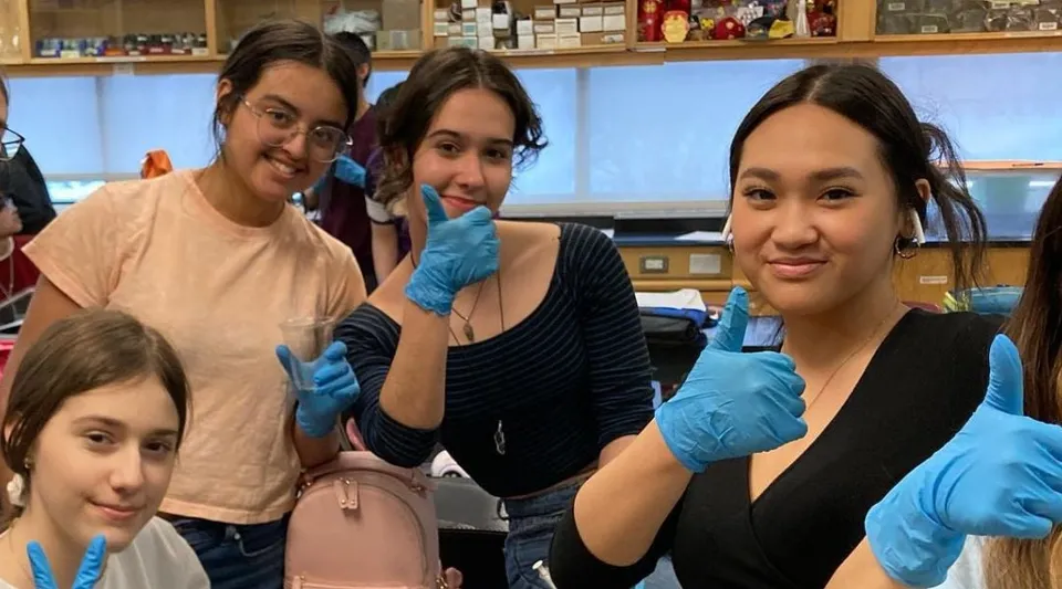 Quattro studentesse del St. James Collegiate indossano guanti e posano insieme in un laboratorio con scaffali e attrezzature.