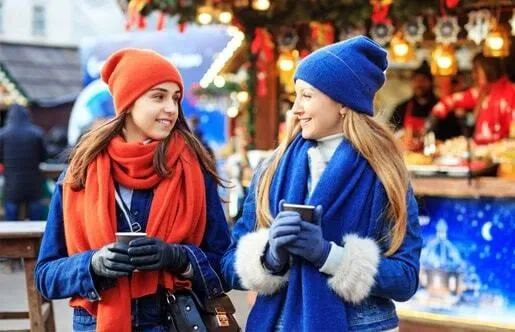 Dos chicas con gorros y bufandas de colores sonríen en un mercado navideño iluminado