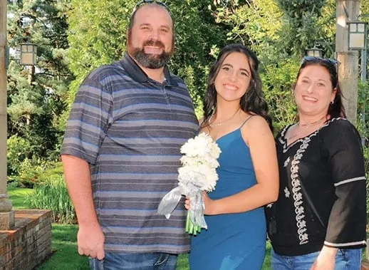 Una joven con vestido azul sostiene un ramo blanco entre un hombre y una mujer