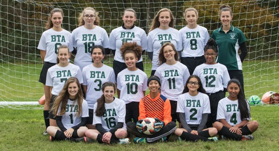 Un gruppo di giovani calciatrici della Boston Trinity Academy posa insieme su un campo d'erba con un pallone da calcio in primo piano.