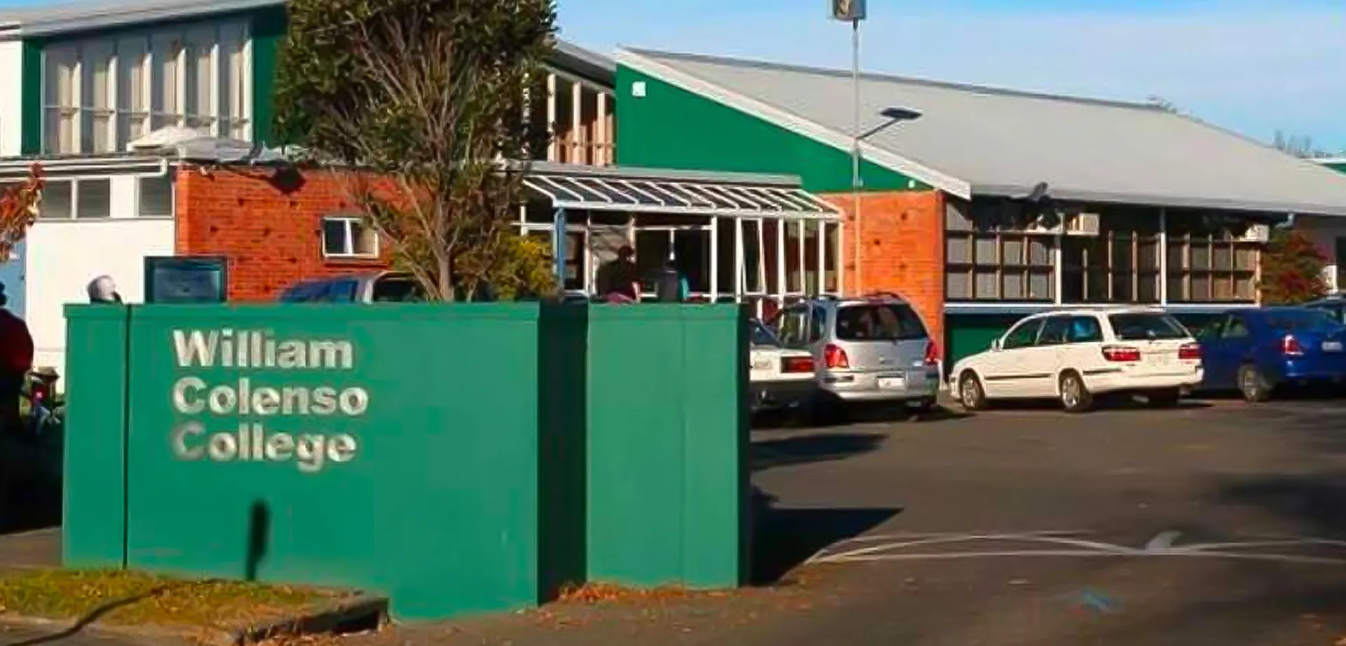 Un cartello verde con il nome William Colenso College davanti a un edificio in mattoni e auto parcheggiate.