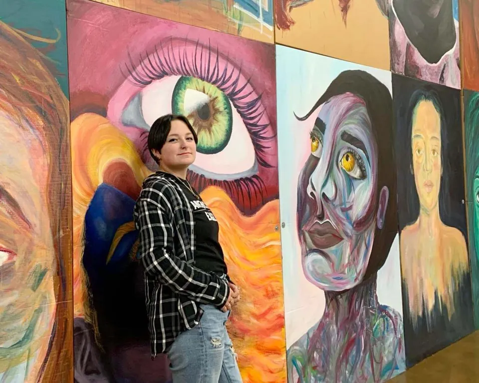 Una persona con una camicia a quadri in piedi davanti a un murale colorato e astratto con forme di occhi surreali nel campus della Mount Lebanon High School.
