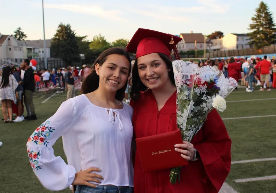 Due studentesse della Mount Everett Regional High School in posa con abiti da diploma davanti a una folla su un campo sportivo.