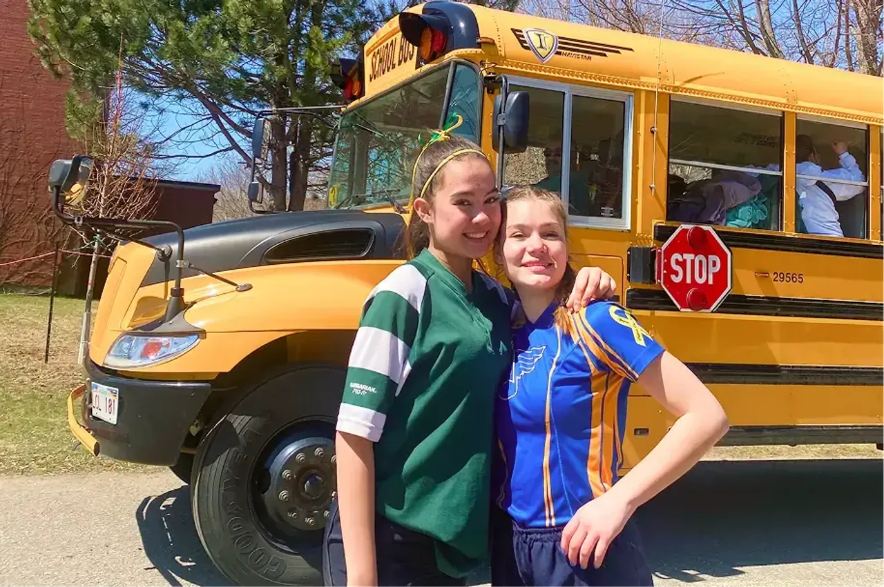 Due studentesse con maglie sportive davanti a uno scuolabus giallo con segnale di stop