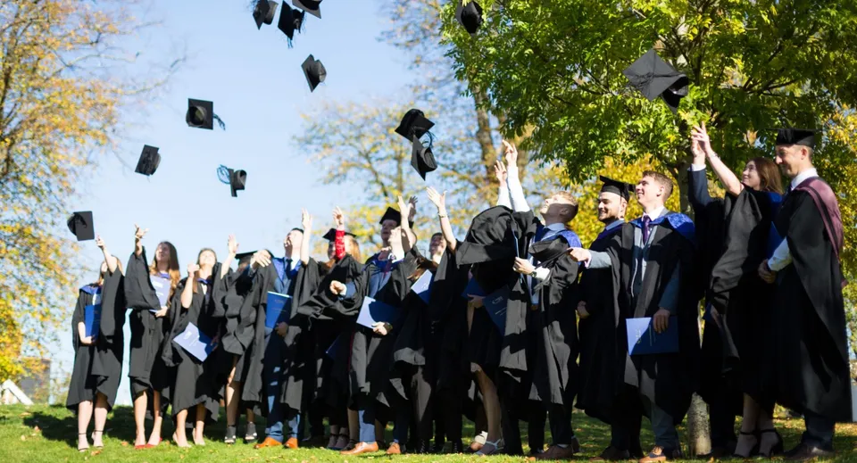 I diplomati del Cheltenham College lanciano in aria i loro tocchi da laurea davanti a verdi alberi.