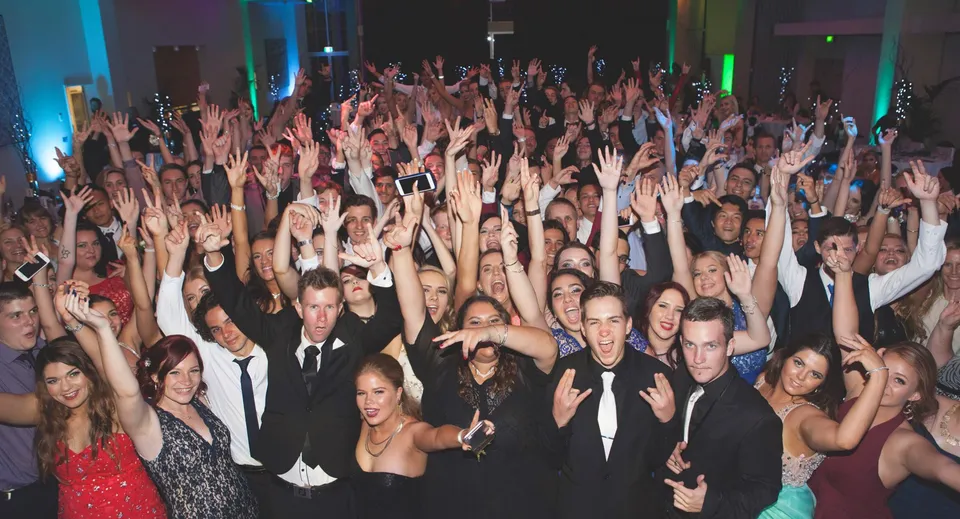 Un grande gruppo di studenti della Ipswich State High School alza le mani in una sala eventi illuminata da luci colorate.