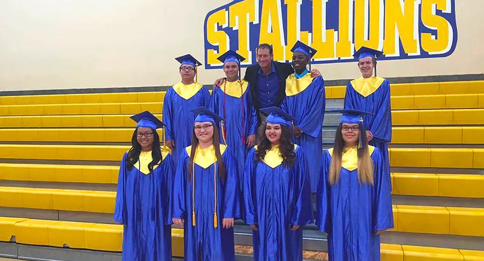 Un gruppo di diplomate e diplomati in toghe blu si trova davanti all'insegna Stallions sulle tribune gialle della Sequoia Charter High School.