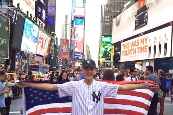 Chico sonriente con gorra y camiseta de béisbol sosteniendo la bandera estadounidense en Times Square
