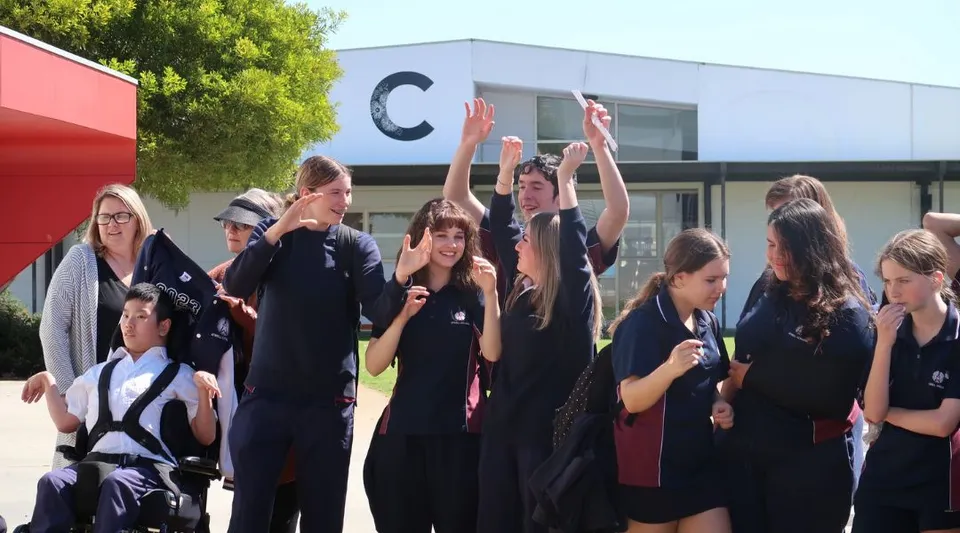 Un gruppo di studenti e studentesse si raccoglie all'aperto davanti a un edificio dell'Atwell College, alcuni alzano le braccia.